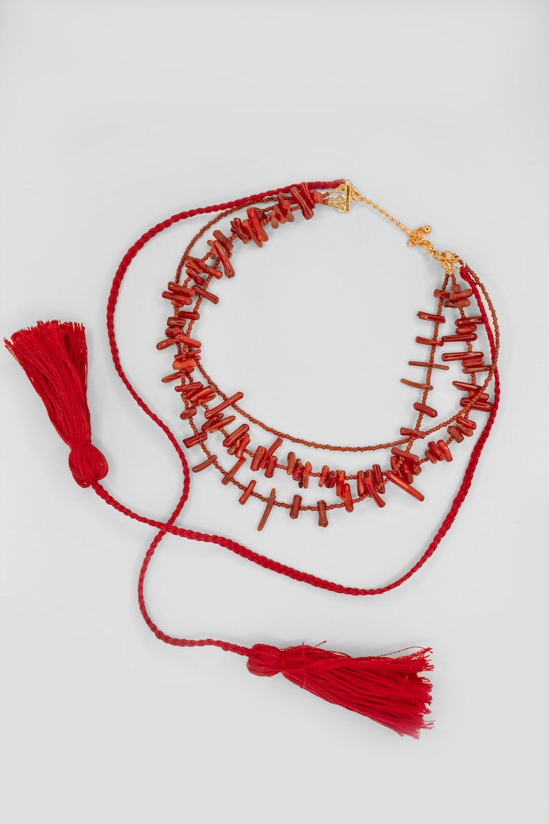 May include: A multi-strand necklace featuring red coral beads and red tassels. The necklace is crafted from braided red cord and has a gold clasp.