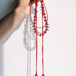 May include: A red coral necklace with two long tassels. The necklace is made of small, irregular pieces of coral that are strung together on a red cord. The tassels are also made of red cord and have a fringe at the bottom.