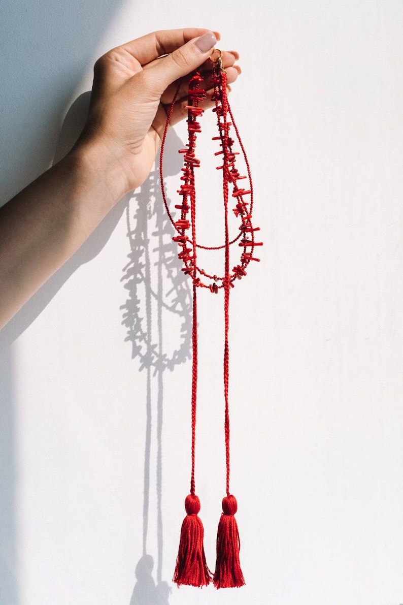 May include: A red coral necklace with two long tassels. The necklace is made of small, irregular pieces of coral that are strung together on a red cord. The tassels are also made of red cord and have a fringe at the bottom.