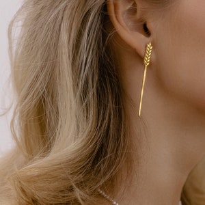Könnte beinhalten: Eine Frau trägt eine zarte Perlenkette mit einem goldenen Weizen-Anhänger. Die Kette ist schlicht und elegant, und der Weizen-Anhänger verleiht ihr einen Hauch von rustikalem Charme.