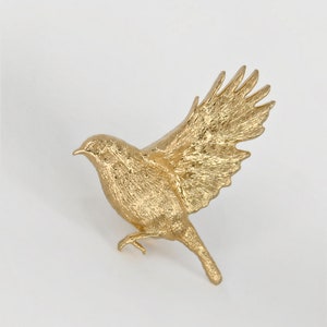 May include: A gold-coloured bird figurine with its wings spread out in flight. The figurine is made of a textured material, giving it a realistic look.
