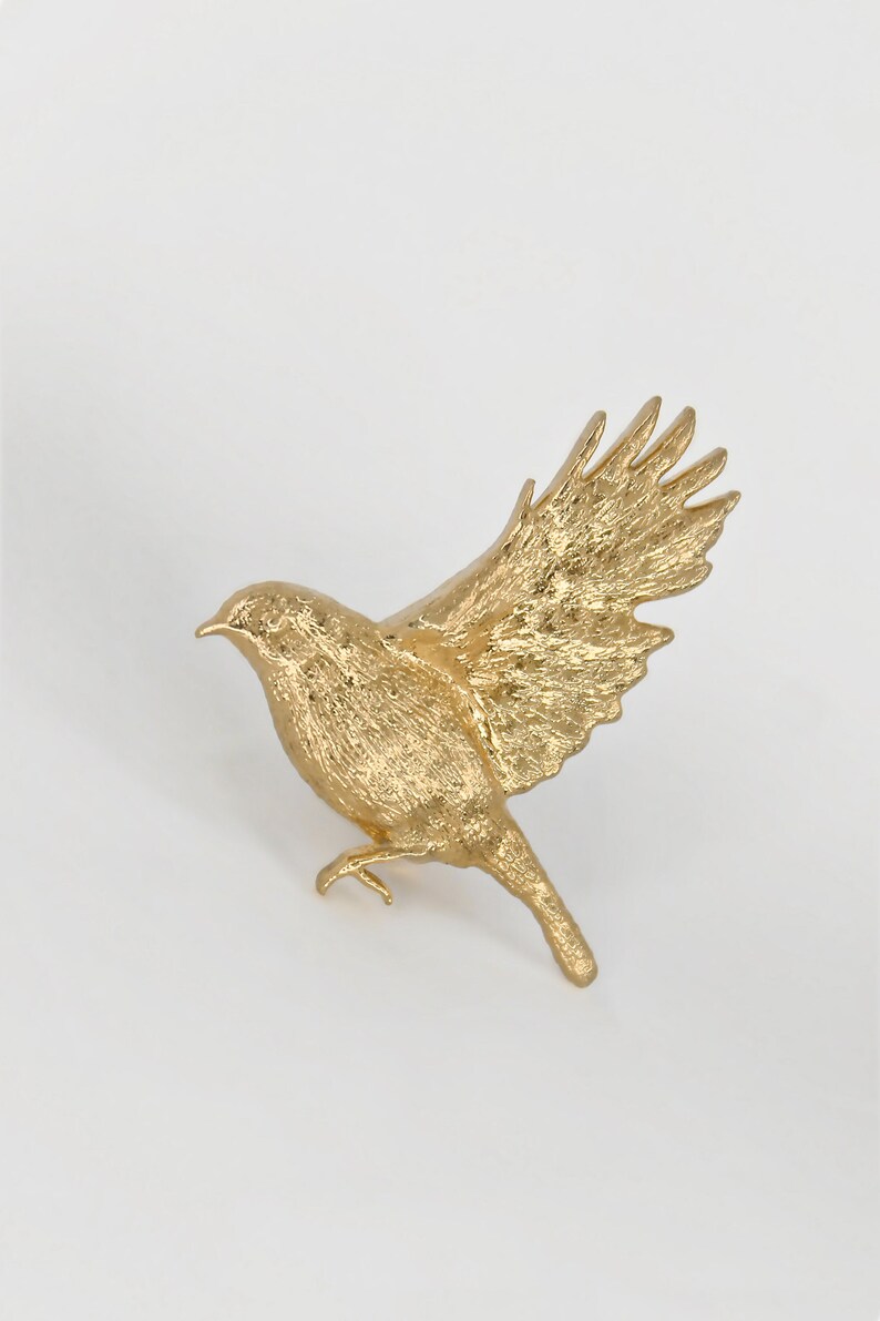 May include: A gold-coloured bird figurine with its wings spread out in flight. The figurine is made of a textured material, giving it a realistic look.