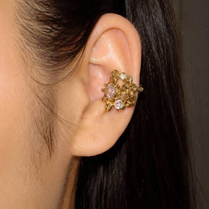 May include: A gold-toned ear cuff featuring an organic, textured metal design with multiple embedded clear, round-cut stones. The piece wraps around the concha area of the ear, showcasing a polished finish and a cluster of sparkling accents.