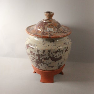 May include: A ceramic jar with a lid, featuring a white glaze with brown and orange speckles. The jar has a round body and a three-legged base. The lid has a label that reads "U-144 B".