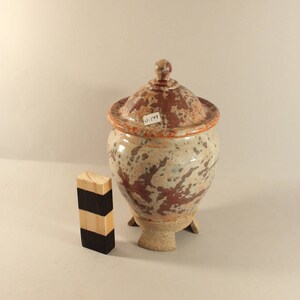 May include: A ceramic jar with a lid, featuring a mottled brown and white glaze. The jar has three small feet and a label that reads "U-144".