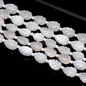 May include: Close-up of several strands of white leaf-shaped beads. The beads are strung together with small, round white beads. The leaves have a frosted appearance and are arranged diagonally against a black background.