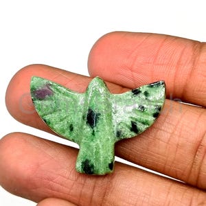 May include: A carved green stone bird figurine with black and red inclusions. The wings are spread wide, and the bird is held in a hand. The stone has a speckled appearance.