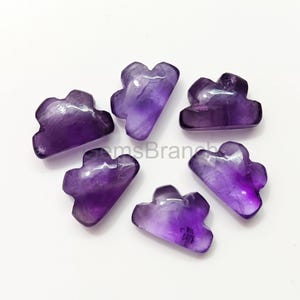 May include: Six amethyst cloud-shaped gemstones. The gemstones are a deep purple colour with varying shades and translucence. The clouds are carved with rounded edges and a smooth finish. The gemstones are arranged on a white surface.