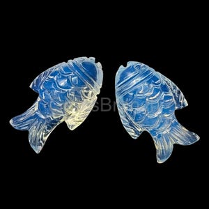 May include: Two opalescent, carved fish figurines. Each fish is detailed with scales and fins, and has a slightly translucent appearance. The background is solid black, highlighting the fish's form and intricate carvings.