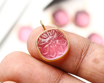 Puede incluir: Un colgante cuadrado de piedra preciosa rosa con un diseño floral tallado en la superficie. El colgante está engastado en un marco de metal dorado y tiene un pequeño lazo en la parte superior para una cadena. El fondo muestra otras piedras preciosas rosas.