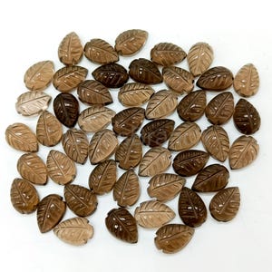 May include: A collection of carved gemstone leaf pendants in varying shades of brown. The leaves are detailed with veining and have a polished finish. The pendants are arranged on a white surface, showcasing their natural colors and textures.