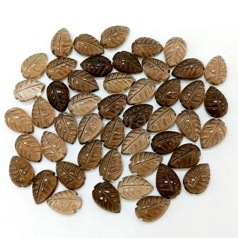 May include: A collection of carved gemstone leaf pendants in varying shades of brown. The leaves are detailed with veining and have a polished finish. The pendants are arranged on a white surface, showcasing their natural colors and textures.