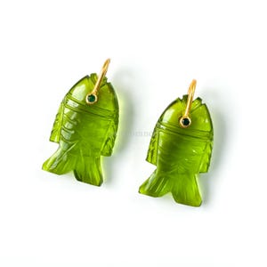 Puede incluir: Un par de pendientes verdes con forma de pez. Cada pendiente presenta un diseño de pez tallado con un gancho dorado y una pequeña gema verde. Los pendientes están sobre un fondo blanco.