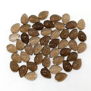 May include: A collection of carved leaf-shaped gemstones in varying shades of brown. The leaves have detailed veining and are arranged on a white surface. The gemstones appear to be polished and have a translucent quality.