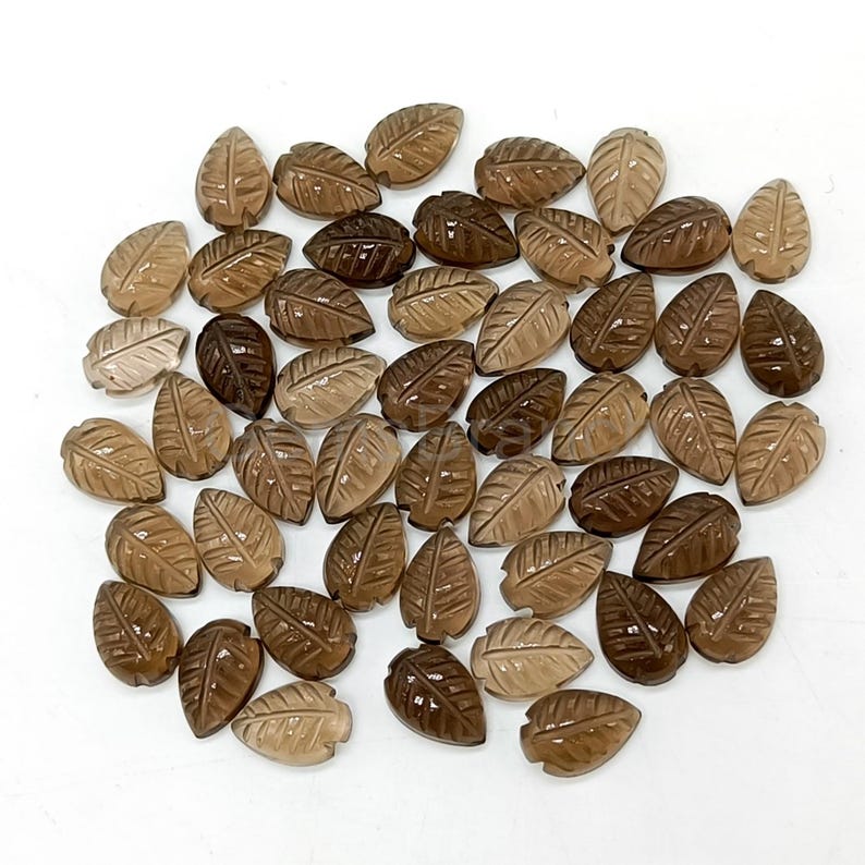 May include: A collection of carved leaf-shaped gemstones in varying shades of brown. The leaves have detailed veining and are arranged on a white surface. The gemstones appear to be polished and have a translucent quality.