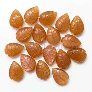 Peut inclure: Collection de pierres précieuses en forme de feuilles, de couleur orange-brun. Chaque gemme est sculptée avec des veines détaillées et a un aspect scintillant et pailleté. Les feuilles sont dispersées sur une surface blanche.