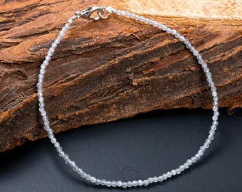 May include: Delicate anklet featuring small, light-colored beads strung together. The beads are evenly spaced, creating a subtle sparkle. The anklet has a silver-colored clasp. The background is a textured, dark brown wood surface.