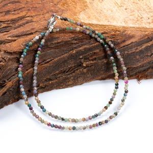 Op de afbeelding: Een delicate ketting met twee strengen kleine, veelkleurige kralen. De kralen vertonen een reeks kleuren, waaronder groen, paars en roze, en zijn aan elkaar geregen met een zilveren sluiting. De ketting wordt gepresenteerd op een getextureerd, bruin houten oppervlak.