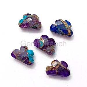 Pode incluir: Cinco contas de pedras preciosas em forma de nuvem em tons de roxo, azul e dourado. As pedras preciosas têm um corte facetado e um acabamento polido. As contas estão dispostas sobre uma superfície branca.