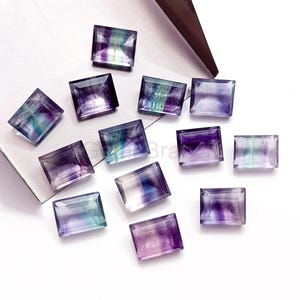 May include: A collection of rectangular fluorite gemstones. The stones display a mix of purple, green, and clear colors. The gemstones are cut with a smooth, polished finish, and are arranged on a white surface.