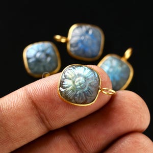 Könnte beinhalten: Vier quadratische Anhänger mit einem floralen Design, das in einen blau-grünen Stein geschnitzt ist. Jeder Anhänger ist in einen goldfarbenen Metallrahmen gefasst und hat eine kleine Schlaufe oben. Der Hintergrund ist schwarz.