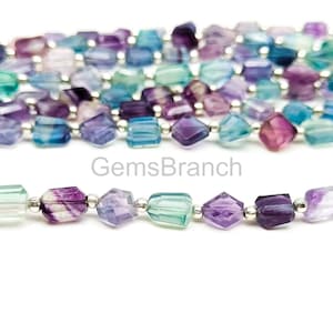May include: Close-up of strands of faceted fluorite beads in shades of purple, blue, and green. The beads are strung on a silver-colored wire. The text "GemsBranch" is visible in the image.