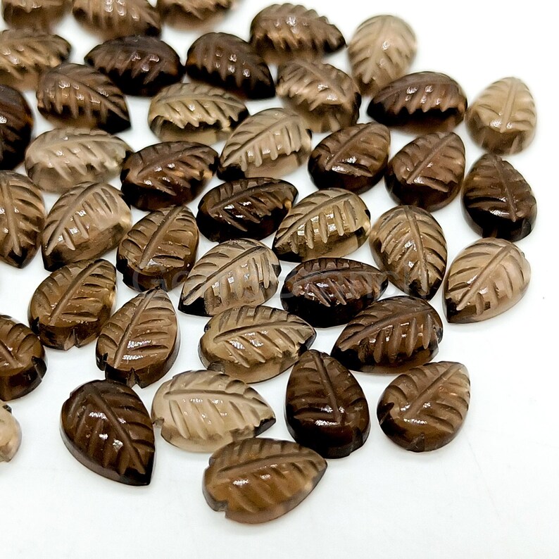 May include: A collection of leaf-shaped gemstones in varying shades of brown. Each gemstone is carved with detailed leaf veins, creating a naturalistic appearance. The gemstones are scattered across a white surface, showcasing their polished finish.