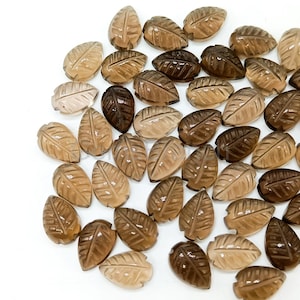 May include: A collection of carved leaf-shaped gemstones in varying shades of brown. The leaves have detailed veining and are scattered on a white surface. The gemstones range in color from light tan to dark brown.