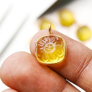 Könnte beinhalten: Ein goldfarbener Anhänger mit quadratischer Form und einem geschnitzten Blumendesign. Der Anhänger hat oben eine Schlaufe für eine Kette. Der Edelstein hat eine durchscheinende gelbe Farbe.
