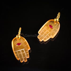 K&ouml;nnte beinhalten: Zwei Anh&auml;nger in Form von H&auml;nden, jeweils mit einem kleinen, ovalen, roten Edelstein in der Mitte. Die Anh&auml;nger sind von transluzenter orangefarbener Farbe mit goldfarbenen Metallakzenten und einer gedrehten Schlaufe zum Aufh&auml;ngen.