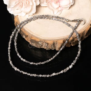 Puede incluir: Un delicado collar compuesto por numerosas cuentas pequeñas, de forma irregular y tono gris. El collar está colocado sobre una superficie de madera, con un fondo negro. El collar se asegura con un cierre plateado. Flores rosas en el fondo.