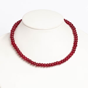 May include: A red beaded necklace displayed on a white mannequin. The necklace is composed of small, round, dark red beads. The beads are strung together to form a single strand necklace. The necklace is a jewelry item.
