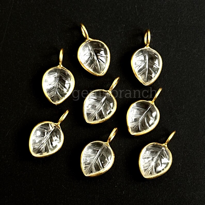 May include: A collection of leaf-shaped pendants. Each pendant features a clear, leaf-veined design encased in a gold-colored metal frame. The pendants are displayed against a black background, highlighting their intricate details.