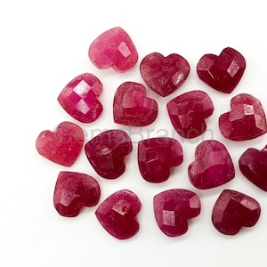 May include: A collection of faceted, heart-shaped gemstones in varying shades of red and pink. The gemstones are scattered on a white surface, each with multiple facets that catch the light, creating a sparkling effect. The hearts vary in size and color intensity.