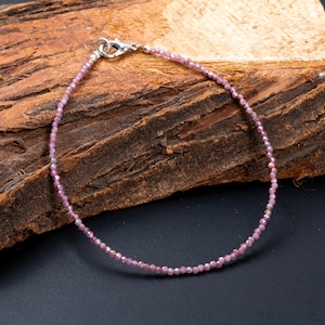 May include: A delicate pink gemstone bracelet with a silver clasp. The bracelet is made of small, round, faceted beads. The bracelet is displayed on a piece of wood with a dark background.