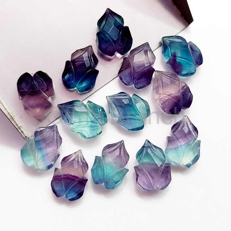 Puede incluir: Una colecci&oacute;n de gemas de fluorita talladas en forma de flores estilizadas. Las gemas muestran una gama de colores, incluyendo tonos de p&uacute;rpura, azul y verde. Las superficies facetadas capturan la luz, creando un efecto brillante. Las gemas est&aacute;n dispuestas sobre una superficie blanca.