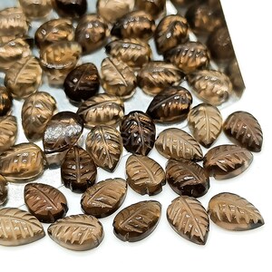 May include: A collection of brown, leaf-shaped gemstones. Each gem is carved with detailed leaf vein patterns. The stones vary in shade from light tan to dark brown. The gemstones are scattered on a white surface, creating a natural, earthy aesthetic.