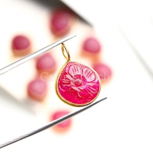 Puede incluir: Un colgante en forma de lágrima con un diseño floral tallado, sostenido por unas pinzas. El colgante es de color rosa vibrante, engastado en un marco dorado. Varias otras gemas rosas están borrosas en el fondo.