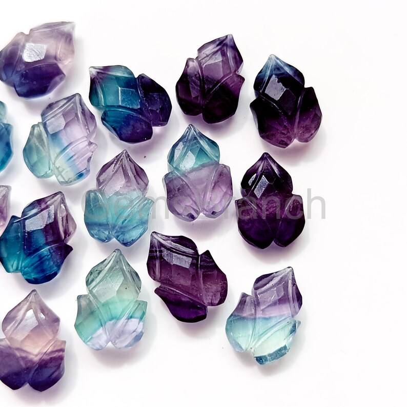 Puede incluir: Una colecci&oacute;n de gemas de fluorita talladas en forma de flores. Las gemas muestran una gama de colores, incluyendo p&uacute;rpura, azul y verde. Las facetas de las piedras capturan la luz, creando un efecto brillante. Las gemas est&aacute;n dispuestas sobre una superficie blanca.