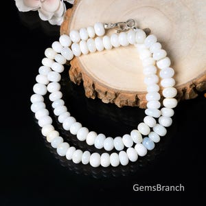 May include: A white beaded necklace with a silver clasp. The necklace is made of small, oval-shaped beads. The necklace is displayed on a wooden surface. The text "GemsBranch" is visible in the bottom right corner.