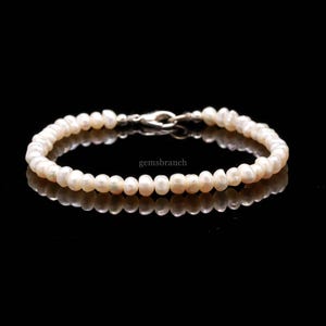 May include: A delicate bracelet composed of small, round, off-white pearls. The pearls are strung together with a silver clasp. The bracelet is displayed on a reflective black surface, with the text "gemsbranch" visible.