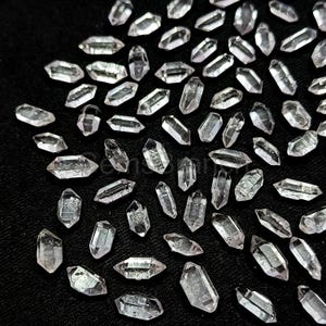 Pode incluir: Uma coleção de pontas de quartzo transparente, também conhecidas como cristais de quartzo duplamente terminados, sobre um fundo preto. As pontas são de vários tamanhos e formas, algumas são mais alongadas do que outras.