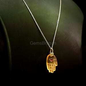 K&ouml;nnte beinhalten: Halskette mit silberner Kette und einem handf&ouml;rmigen Anh&auml;nger. Der Anh&auml;nger ist bernsteinfarben und durchscheinend, mit einem kleinen roten Edelstein. Die Kette wird vor dunklem Hintergrund pr&auml;sentiert.