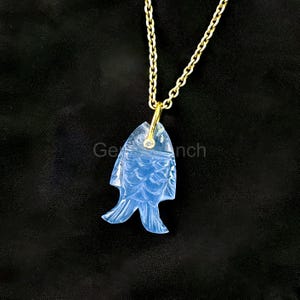 May include: A light blue, fish-shaped pendant necklace with a gold chain. The pendant is carved with detailed scales and fins, and a small diamond is set near the top. The necklace hangs against a black background.