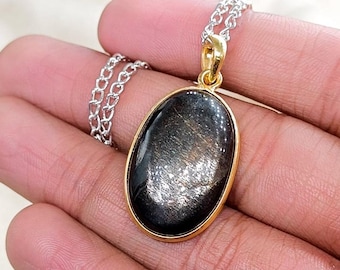 May include: A gold-toned pendant necklace with an oval, black gemstone. The gemstone has a shimmering, reflective quality. The chain is silver-colored and delicate. The pendant is held in a hand.