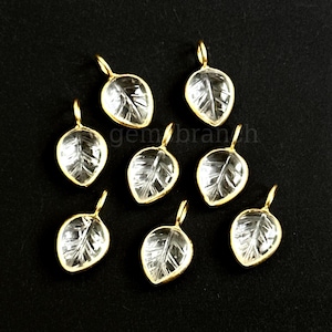 May include: Seven leaf-shaped pendants with a clear, textured center and a gold-colored border. Each pendant has a small loop at the top for attaching to a chain or other jewelry component. The pendants are arranged on a black surface.