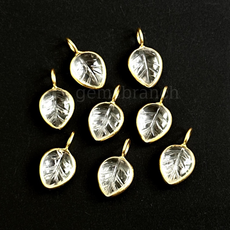May include: Seven leaf-shaped pendants with a clear, textured center and a gold-colored border. Each pendant has a small loop at the top for attaching to a chain or other jewelry component. The pendants are arranged on a black surface.