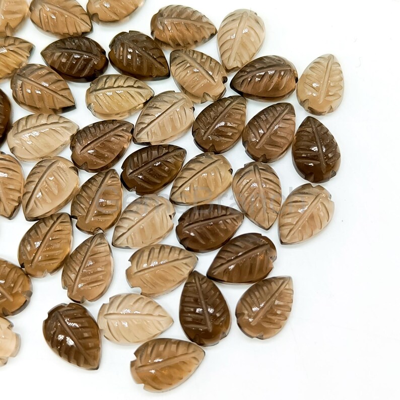 May include: A collection of leaf-shaped gemstones in varying shades of brown, from light tan to deep chocolate. Each gemstone is carved with detailed leaf vein patterns, creating a natural and organic aesthetic. The gemstones are scattered on a white surface.