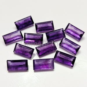 Puede incluir: Una colección de piedras preciosas de amatista rectangulares. Cada gema es de color púrpura intenso con una superficie pulida. Las gemas están dispersas sobre una superficie blanca, mostrando su claridad y color.