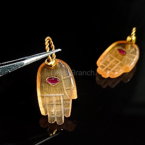 K&ouml;nnte beinhalten: Zwei Anh&auml;nger in Form einer Hand, gefertigt aus einem durchscheinenden, orangefarbenen Material. Jeder Anh&auml;nger hat einen kleinen, ovalen roten Stein. Die Anh&auml;nger haben goldfarbene Metallakzente und sind an einer gedrehten, goldfarbenen Schlaufe aufgeh&auml;ngt.